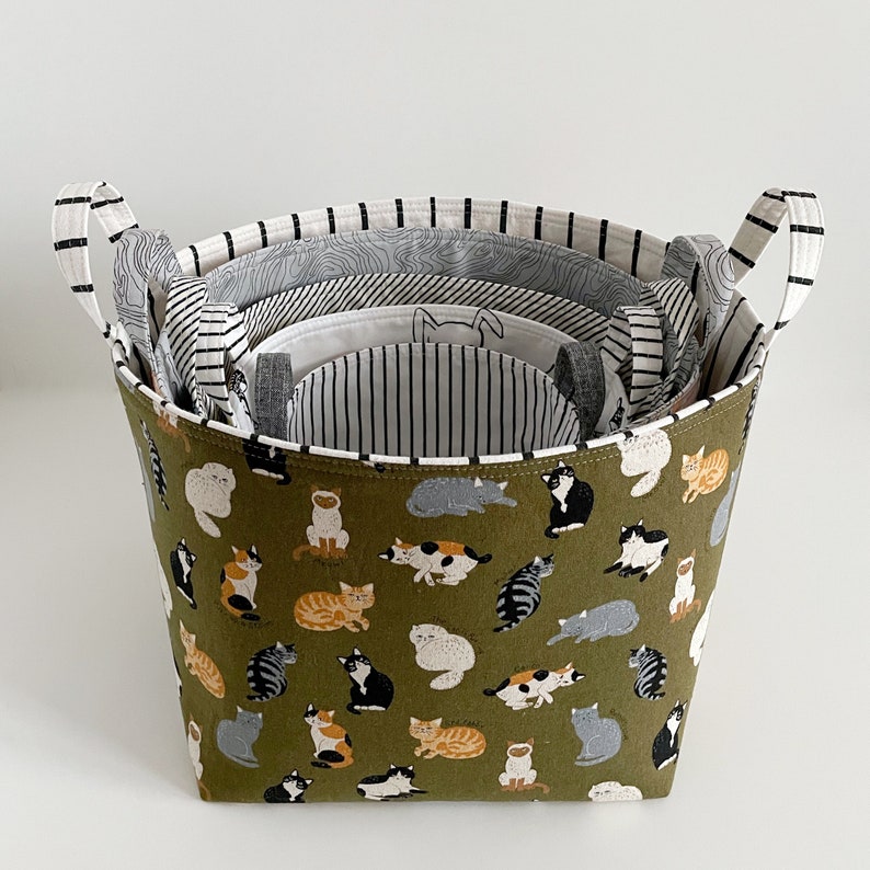 May include: A green fabric storage basket with a cat print design. The basket is made of multiple layers of fabric, with the top layer featuring a green background and a white cat print. The bottom layers are white with black stripes.