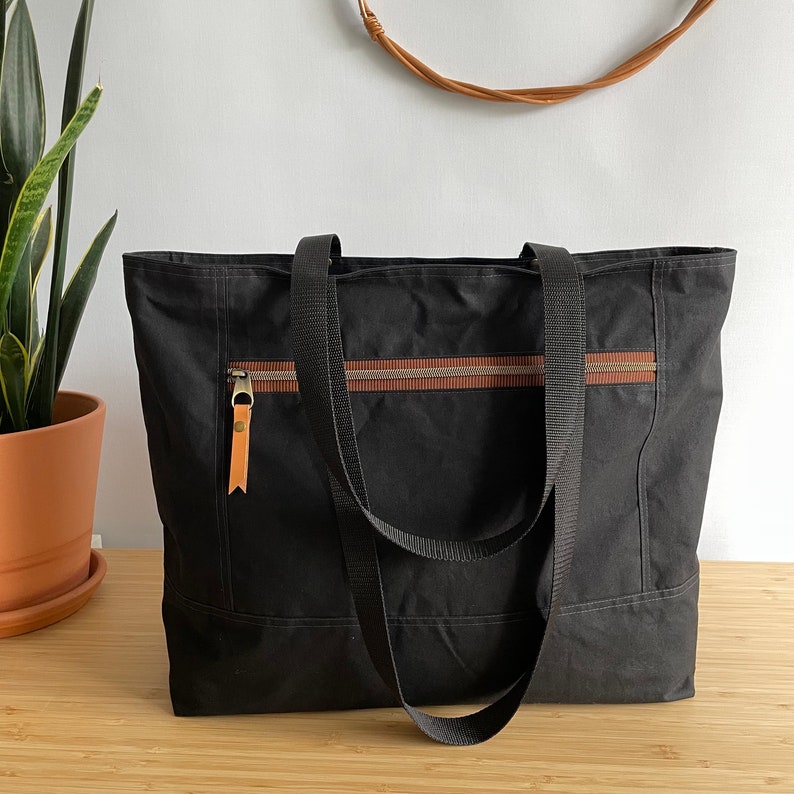 May include: A black canvas tote bag with brown leather accents and a zipper closure. The bag has two long straps and a smaller pocket on the front.