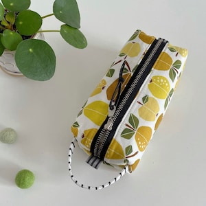 May include: A white zippered pouch with a black zipper and a black and white striped handle. The pouch is decorated with a pattern of yellow pears and green leaves.