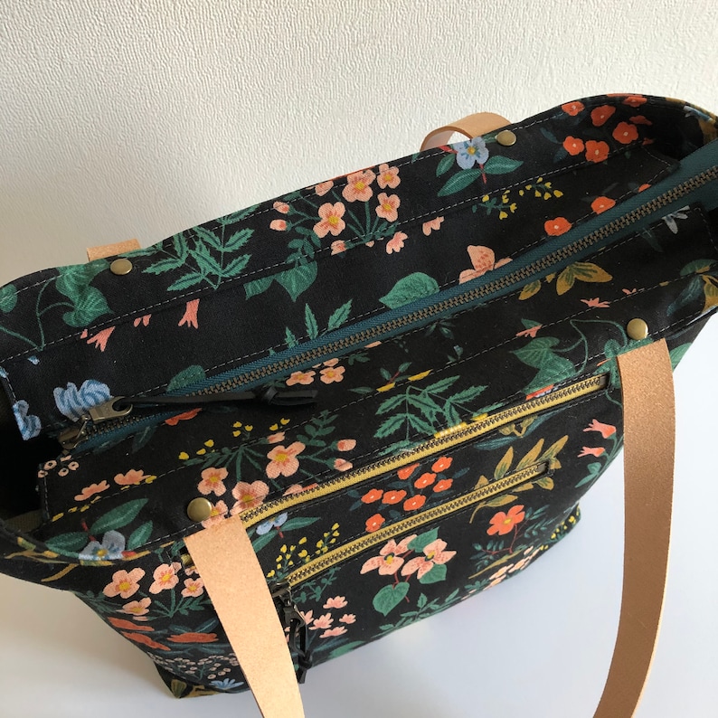 Pode incluir: Uma bolsa tote preta com estampa floral e dois bolsos com z&iacute;per. A bolsa tem al&ccedil;as de couro marrom e um z&iacute;per turquesa.