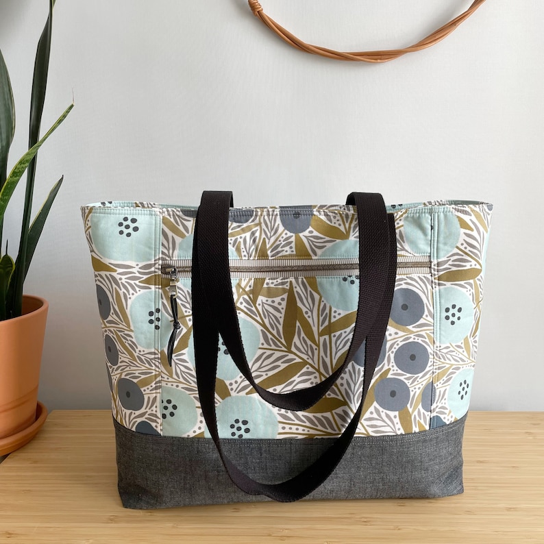 May include: A large tote bag with black straps and a zipped pocket. The bag is made of a light blue fabric with a pattern of brown leaves and blue and white flowers.