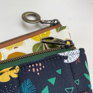 Puede incluir: Una bolsa negra con cremallera, con una cremallera verde y un estampado floral blanco y amarillo. La bolsa tiene una cremallera marr&oacute;n con un tirador de anillo de lat&oacute;n.