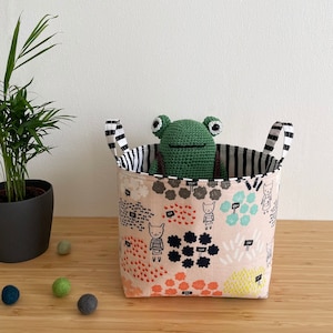 May include: A green crocheted frog toy sits inside a beige and black striped fabric storage bin with a whimsical pattern of flowers, animals, and text.
