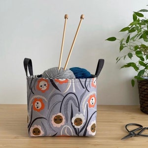 May include: A gray fabric storage basket with black handles and a floral pattern featuring orange flowers with faces. The basket is filled with two balls of yarn and two wooden knitting needles.