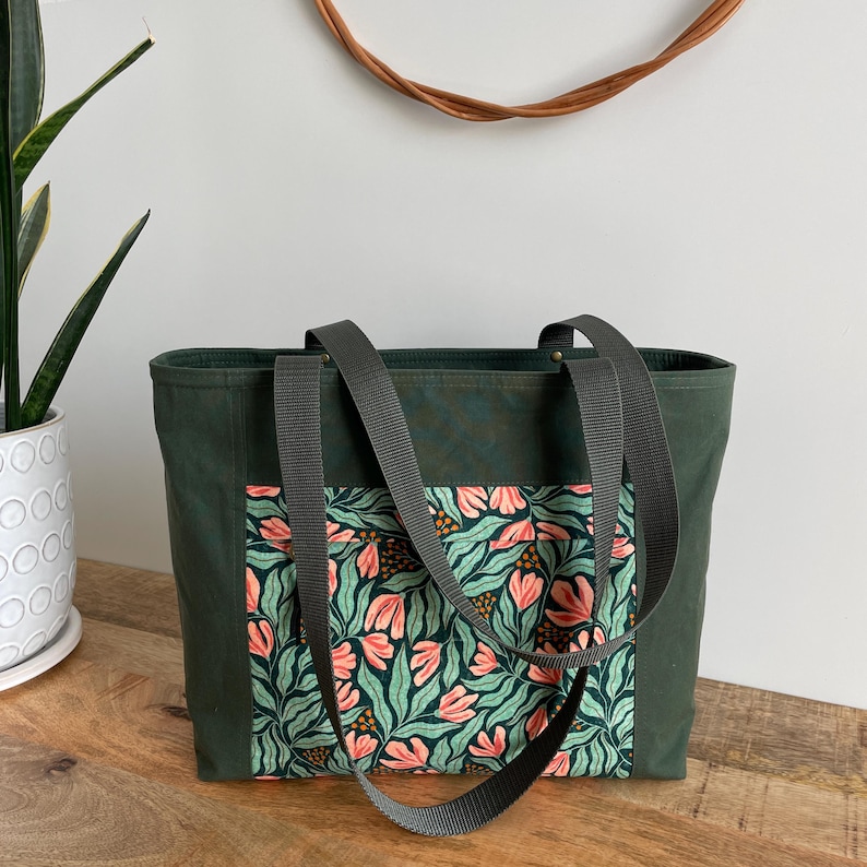 May include: A dark green canvas tote bag with a floral print pocket. The bag has two long straps and a zipper closure.