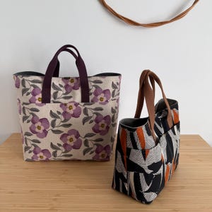May include: Two tote bags. One is beige with a purple floral pattern and dark purple handles. The other is black and white with orange accents and brown handles. Both bags have pockets and are on a wooden surface.
