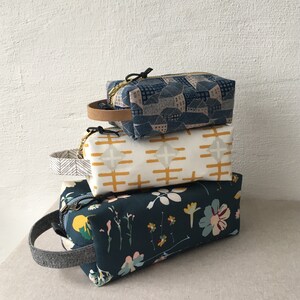 May include: Three fabric zippered pouches stacked on top of each other. The top pouch is blue with a white and pink geometric pattern. The middle pouch is white with a yellow geometric pattern. The bottom pouch is dark blue with a floral pattern.