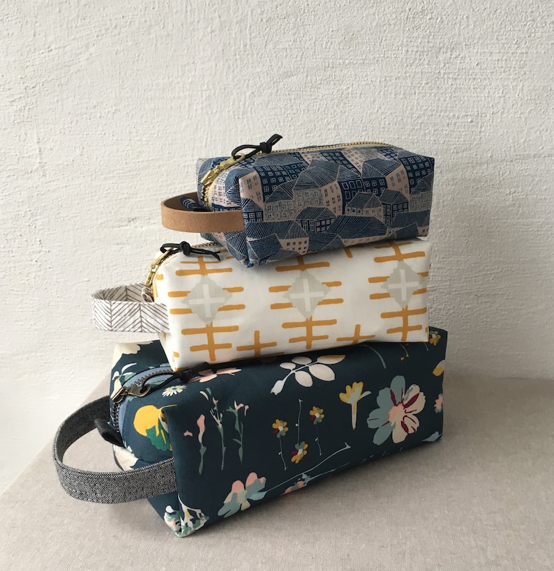 May include: Three fabric zippered pouches stacked on top of each other. The top pouch is blue with a white and pink geometric pattern. The middle pouch is white with a yellow geometric pattern. The bottom pouch is dark blue with a floral pattern.