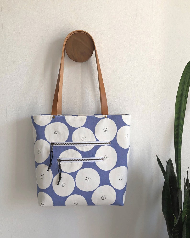Pode incluir: Uma bolsa tote de lona azul com c&iacute;rculos brancos e uma al&ccedil;a de couro marrom. A bolsa tem dois bolsos com z&iacute;per na frente.