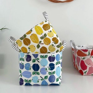 May include: Three fabric storage baskets with different fruit patterns. The top basket has a yellow and white pattern of apples, the middle basket has a blue and white pattern of apples, and the bottom basket has a red and white pattern of pears.