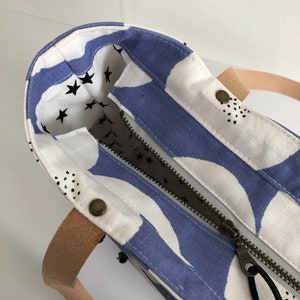Pode incluir: Uma bolsa tote branca com um design estampado em azul e branco. A bolsa tem um fecho de correr e uma al&ccedil;a de couro castanho. O interior da bolsa &eacute; forrado com um tecido branco com estrelas pretas.