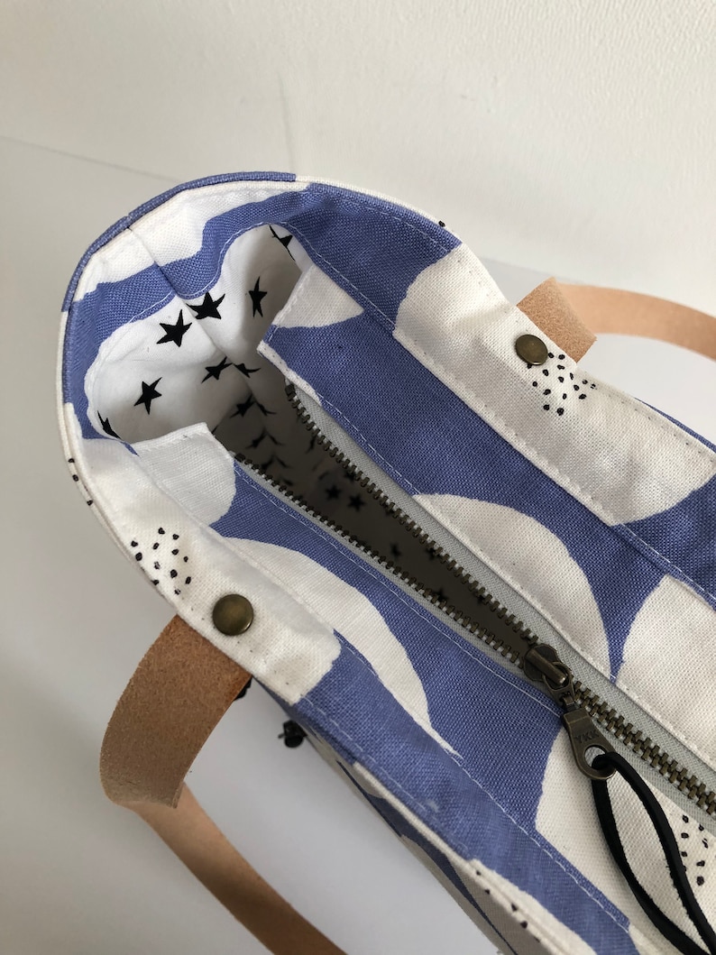 Pode incluir: Uma bolsa tote branca com um design estampado em azul e branco. A bolsa tem um fecho de correr e uma al&ccedil;a de couro castanho. O interior da bolsa &eacute; forrado com um tecido branco com estrelas pretas.