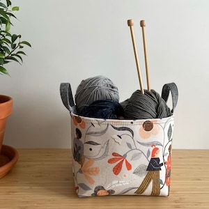 May include: A white fabric storage basket with floral and human figure patterns. The basket has two grey yarn balls and two wooden knitting needles inside.