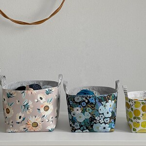 May include: Six fabric storage baskets with different patterns. The baskets are lined up on a white shelf. The patterns include cats, flowers, lemons, and a geometric design.