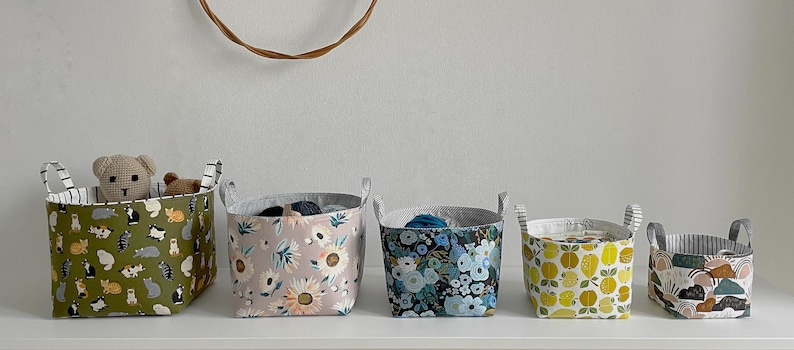 May include: Six fabric storage baskets with different patterns. The baskets are lined up on a white shelf. The patterns include cats, flowers, lemons, and a geometric design.