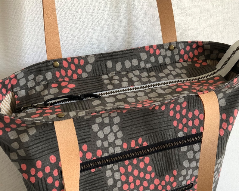 Pode incluir: Uma bolsa tote cinza e rosa com al&ccedil;as de couro e fecho de correr. A bolsa tem um padr&atilde;o geom&eacute;trico com pequenos c&iacute;rculos rosa.