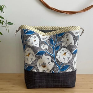 Pode incluir: Uma bolsa de cord&atilde;o com um padr&atilde;o floral branco sobre um fundo azul. A bolsa tem um fundo preto e uma parte superior listrada amarela e branca.