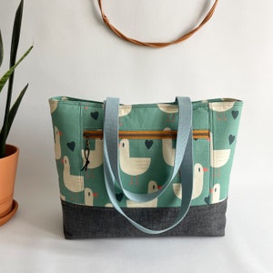 May include: A teal blue tote bag with a zipped pocket and two long straps. The bag is made of a fabric with a pattern of white seagulls and brown hearts. The bottom of the bag is made of a dark denim fabric.