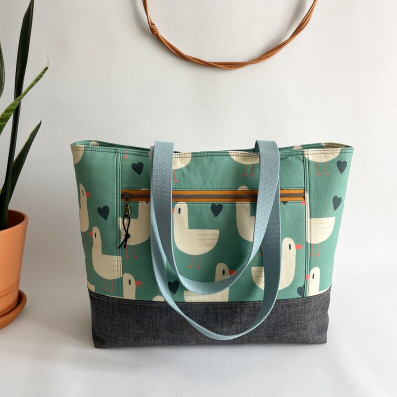 May include: A teal blue tote bag with a zipped pocket and two long straps. The bag is made of a fabric with a pattern of white seagulls and brown hearts. The bottom of the bag is made of a dark denim fabric.
