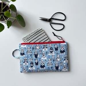 May include: A blue zippered pouch with a repeating pattern of pirates and nautical elements. The pouch is made of quilted fabric and has a red zipper.