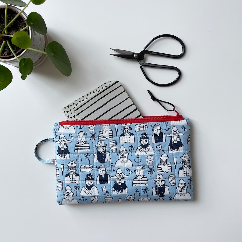May include: A blue zippered pouch with a repeating pattern of pirates and nautical elements. The pouch is made of quilted fabric and has a red zipper.