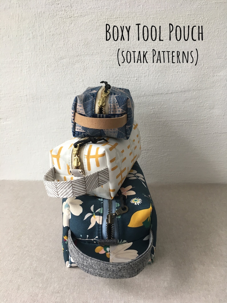 May include: Three stacked fabric pouches with zipper closures and handles. The top pouch is blue with a white geometric pattern, the middle pouch is white with a yellow geometric pattern, and the bottom pouch is blue with a floral pattern. The pouches are labeled "Boxy Tool Pouch (Sotak Patterns)".
