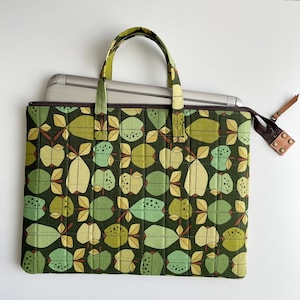Puede incluir: Una bolsa para port&aacute;til verde con cierre de cremallera y una correa de cuero marr&oacute;n. La bolsa tiene un patr&oacute;n repetido de manzanas verdes, marrones y amarillas.