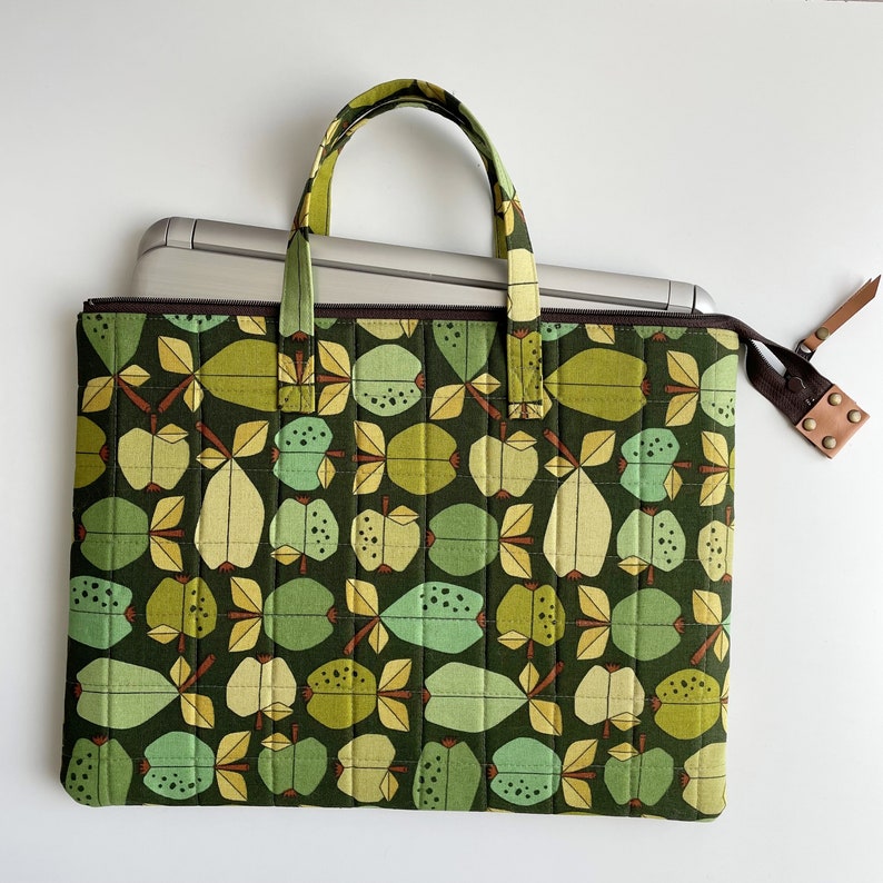 Puede incluir: Una bolsa para port&aacute;til verde con cierre de cremallera y una correa de cuero marr&oacute;n. La bolsa tiene un patr&oacute;n repetido de manzanas verdes, marrones y amarillas.