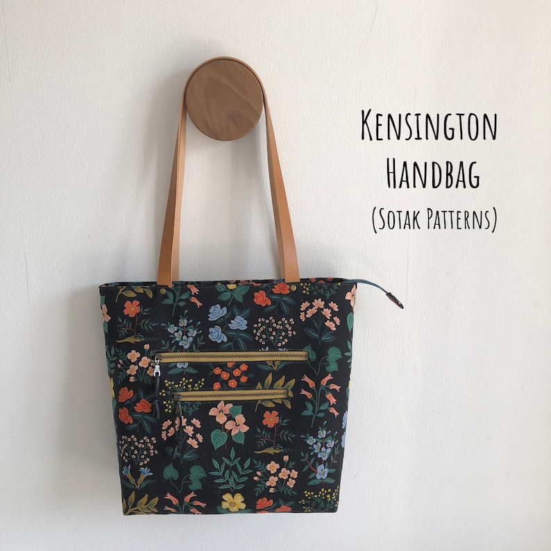 Pode incluir: Uma bolsa de m&atilde;o preta com estampa floral e al&ccedil;as de couro marrom. A bolsa tem dois bolsos com z&iacute;per e o texto "KENSINGTON HANDBAG (SOTAK PATTERNS)" &eacute; vis&iacute;vel.