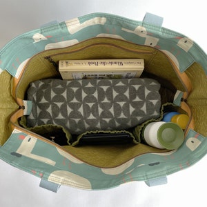 May include: A teal blue tote bag with a white and orange pattern of birds. The bag is open and filled with items, including a book titled "Winnie-the-Pooh", a green and white patterned blanket, a black phone case, and two green and white bottles.