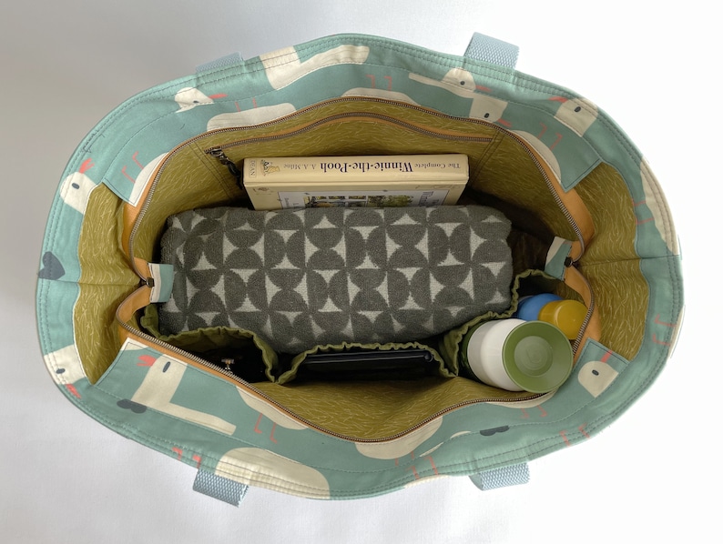 May include: A teal blue tote bag with a white and orange pattern of birds. The bag is open and filled with items, including a book titled "Winnie-the-Pooh", a green and white patterned blanket, a black phone case, and two green and white bottles.