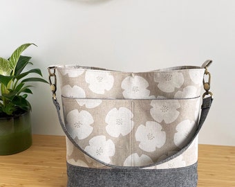 May include: A beige canvas tote bag with a gray bottom and a white floral pattern. The bag has a long adjustable strap and a top zipper closure.