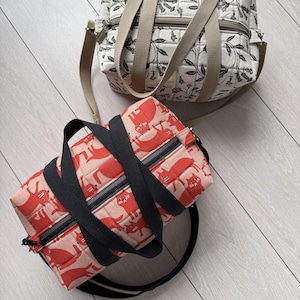 Könnte beinhalten: Zwei gesteppte Reisetaschen mit Reißverschlüssen und Griffen. Eine Tasche ist cremefarben mit schwarzem Blumenmuster und beigen Riemen. Die andere Tasche ist hellrosa mit rotem Katzenmuster und schwarzen Riemen sowie einem schwarzen Schultergurt.