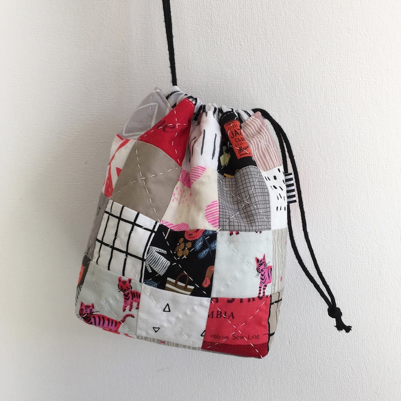 Pode incluir: Uma bolsa de cord&atilde;o de patchwork com uma variedade de tecidos coloridos, incluindo vermelho, rosa, preto, branco e cinza. A bolsa apresenta um padr&atilde;o geom&eacute;trico e uma pequena estampa de um tigre. A bolsa tem um cord&atilde;o de ajuste preto e &eacute; feita de tecido acolchoado.