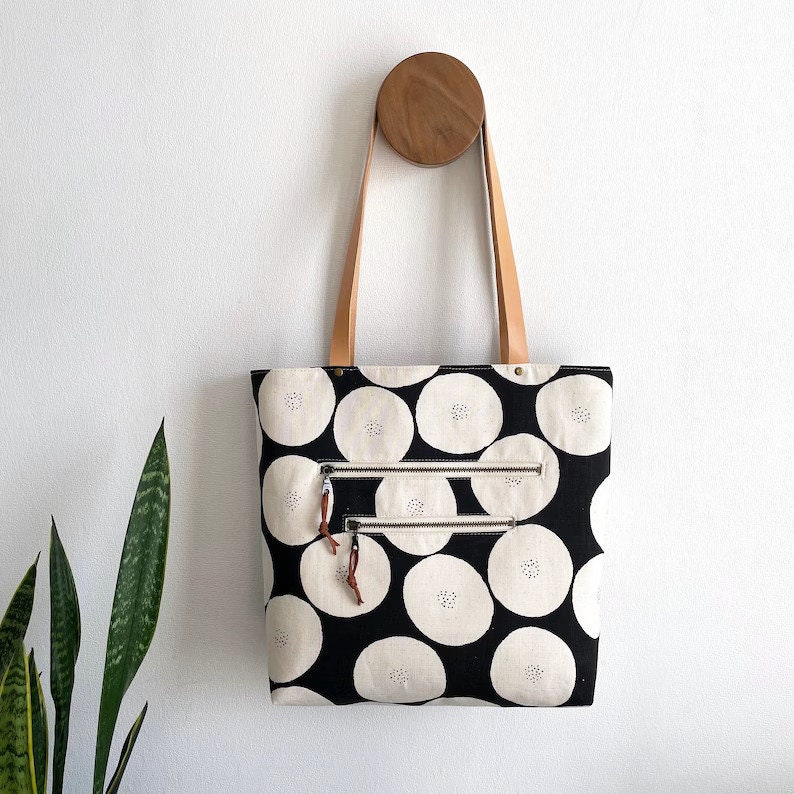 Pode incluir: Uma bolsa tote preta e branca com um padr&atilde;o de c&iacute;rculos. A bolsa tem dois bolsos com z&iacute;per e al&ccedil;as de couro marrom.