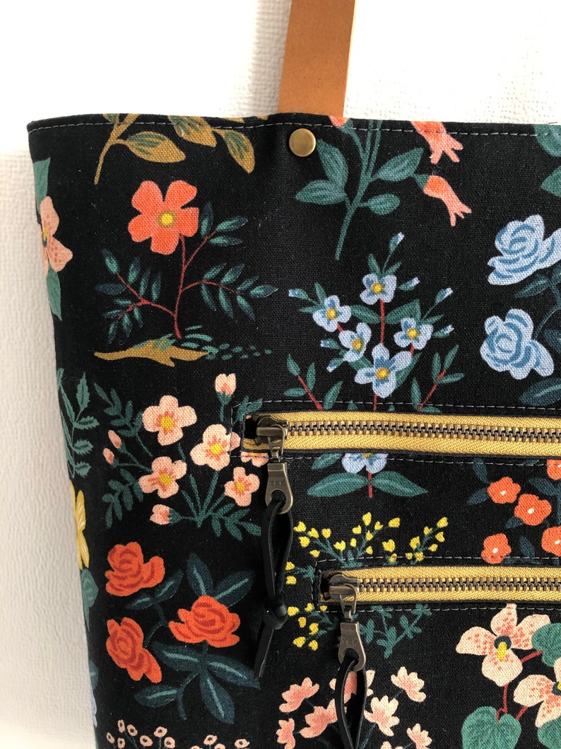 Pode incluir: Sacola tote de lona preta com estampa floral com flores rosa, vermelhas e azuis. A sacola tem dois bolsos com z&iacute;per com z&iacute;peres dourados e puxadores pretos.