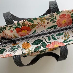 May include: A floral print tote bag with a zipper closure and two black handles. The bag is made of a light-colored fabric with a floral pattern in shades of pink, red, yellow, and green.
