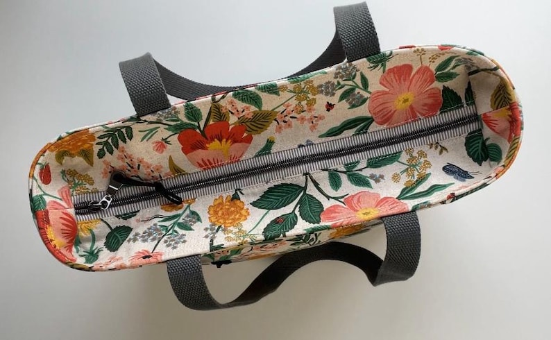 May include: A floral print tote bag with a zipper closure and two black handles. The bag is made of a light-colored fabric with a floral pattern in shades of pink, red, yellow, and green.