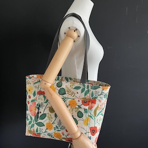May include: A floral tote bag with a white background and a variety of colorful flowers, including red, yellow, and green. The bag has a black strap and a zippered pocket on the front.
