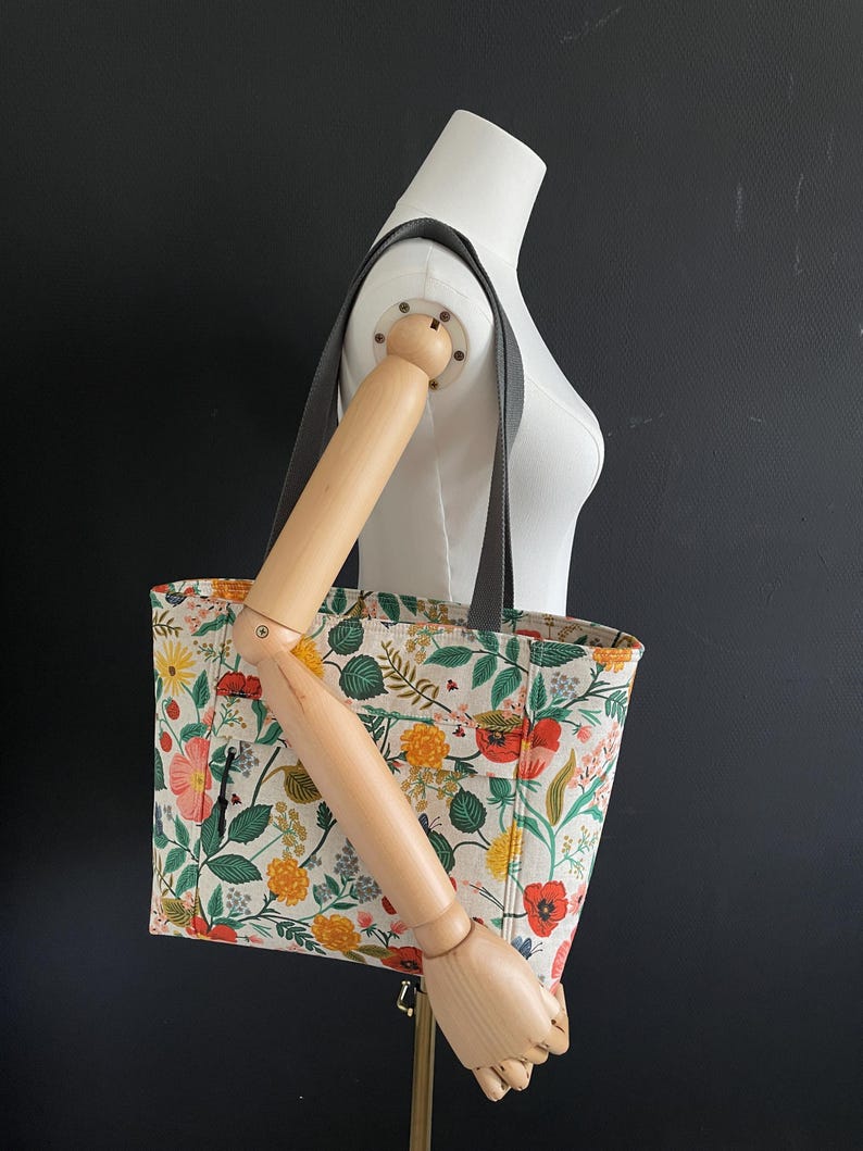 May include: A floral tote bag with a white background and a variety of colorful flowers, including red, yellow, and green. The bag has a black strap and a zippered pocket on the front.