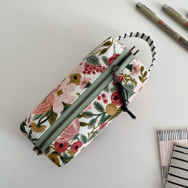 May include: A floral print zippered pencil case with a green zipper and a black zipper pull. The case has a white background with pink, red, and green flowers.