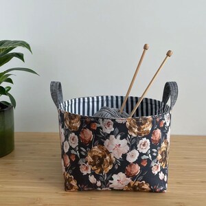 May include: A black and white striped fabric storage basket with a floral print. The basket is filled with a ball of yarn and two wooden knitting needles.