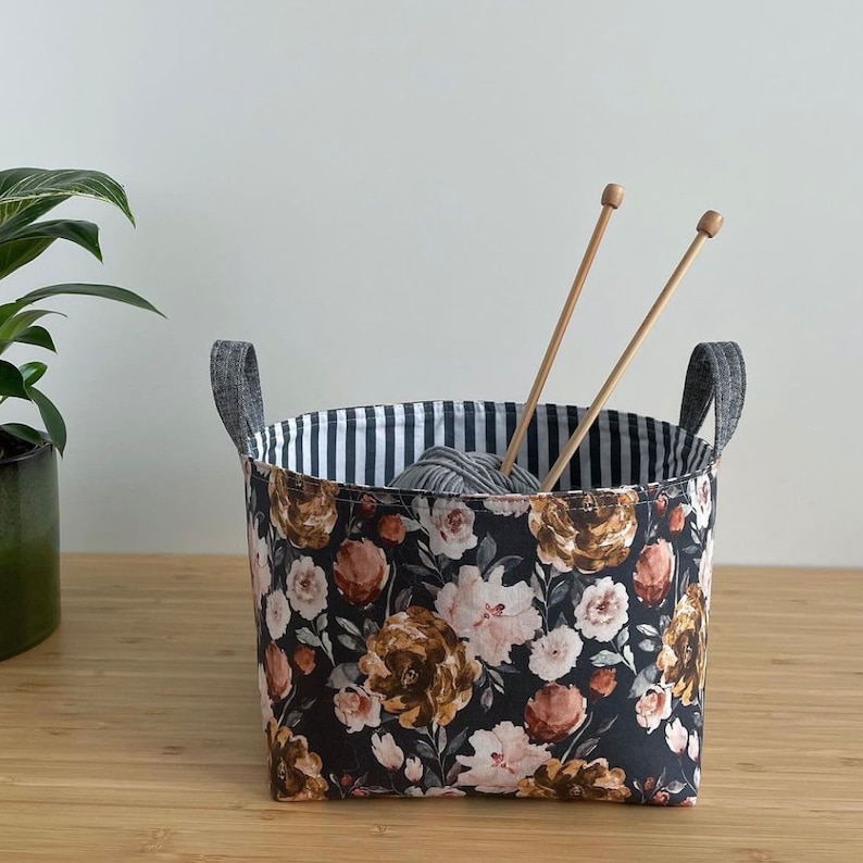 May include: A black and white striped fabric storage basket with a floral print. The basket is filled with a ball of yarn and two wooden knitting needles.