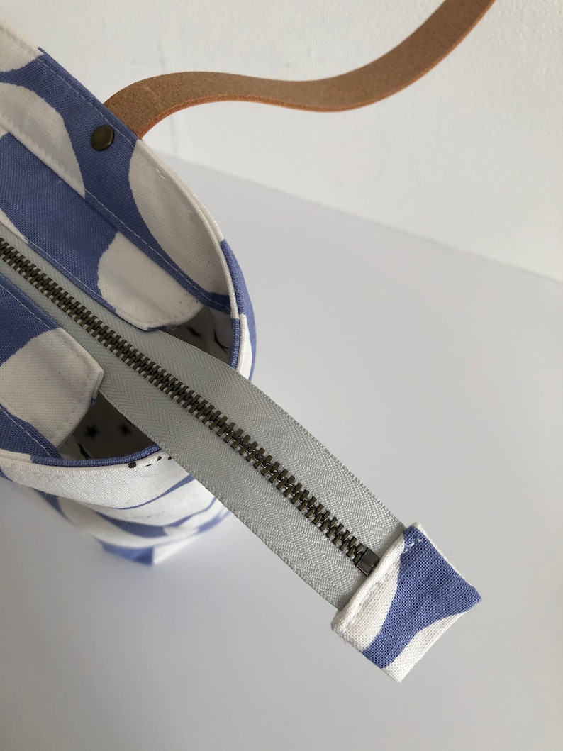 Pode incluir: Uma bolsa tote branca e azul com fecho de correr e al&ccedil;a de couro castanho. A bolsa tem um padr&atilde;o de formas abstratas.