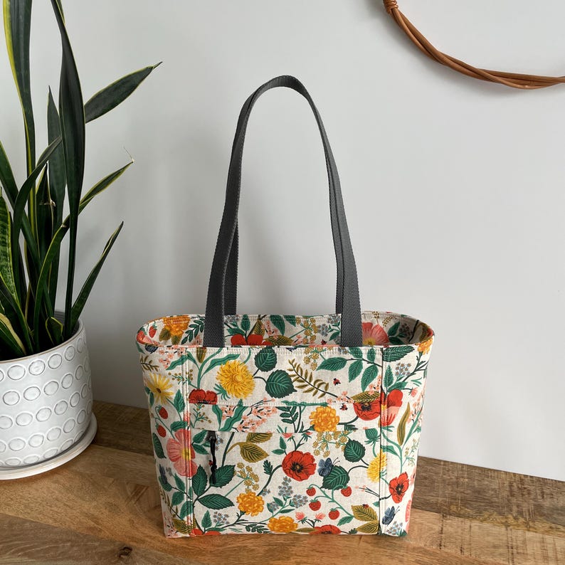 May include: A white tote bag with a floral print featuring red, yellow, and green flowers. The bag has gray handles and a zippered pocket on the front.