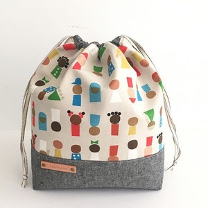 Pode incluir: Uma bolsa de cord&atilde;o feita de tecido branco com fundo cinza. A bolsa tem um padr&atilde;o de pessoas coloridas nela. A bolsa tem uma etiqueta de couro marrom que diz "feito &agrave; m&atilde;o".