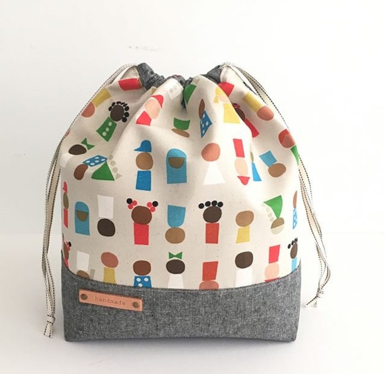 Pode incluir: Uma bolsa de cord&atilde;o feita de tecido branco com fundo cinza. A bolsa tem um padr&atilde;o de pessoas coloridas nela. A bolsa tem uma etiqueta de couro marrom que diz "feito &agrave; m&atilde;o".