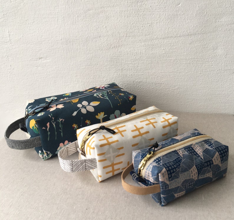 May include: Three zippered pouches with different fabric patterns. The largest pouch is navy blue with a floral pattern, the middle pouch is white with a yellow geometric pattern, and the smallest pouch is blue with a geometric pattern.