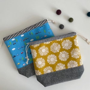 Pode incluir: Duas bolsas de cord&atilde;o com padr&otilde;es diferentes. Uma bolsa &eacute; azul com uma faixa branca e um padr&atilde;o de carros e nuvens. A outra bolsa &eacute; amarela com um padr&atilde;o de raios de sol brancos. Ambas as bolsas t&ecirc;m um fundo cinzento.