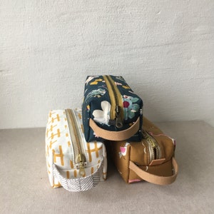 May include: Three zippered pouches with fabric handles. The pouch on top has a floral pattern in shades of blue, green, and white. The middle pouch has a yellow and white geometric pattern. The bottom pouch has a brown and white geometric pattern.